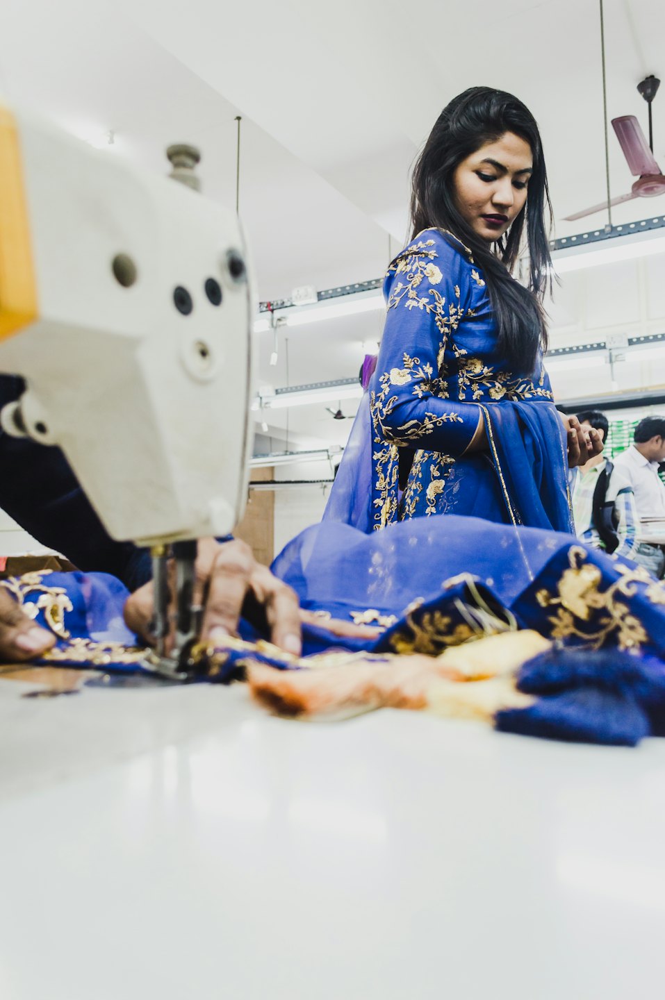 Woman in blue dress watches hands sew gold pattern on fabric.